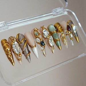 May include: A set of ten long, almond-shaped press-on nails in a clear plastic case. The nails feature a variety of designs, including gold embellishments, rhinestones, and glitter accents. Colors include gold, silver, clear, and various shades of green and pink.