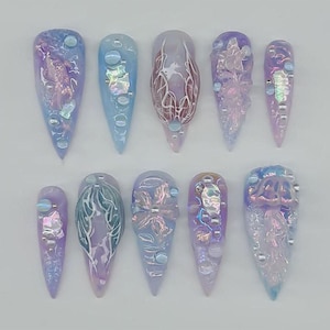May include: A set of ten long, pointed artificial nails. Each nail is a different design, featuring a mix of pastel colors like lavender, blue, and pink. Some nails have iridescent accents, while others have intricate, raised patterns and small, round embellishments.