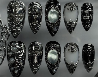 Könnte beinhalten: Ein Set aus zehn künstlichen Nägeln im Gothic-Stil. Die Nägel weisen komplizierte silberne und schwarze Designs auf, darunter Kreuze, ovale Akzente und Perlenverzierungen. Die Nägel sind mandelförmig und haben ein glänzendes Finish.