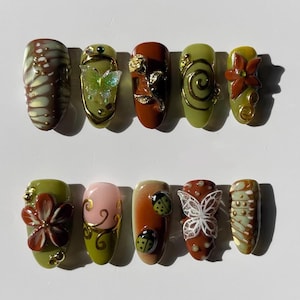Könnte beinhalten: Ein Set aus zehn künstlichen Nägeln mit verschiedenen Designs. Die Nägel zeigen Farben wie Braun, Grün und Rosa, mit Verzierungen wie Schmetterlingen, Blumen und Wirbeln. Einige Nägel haben ein glänzendes Finish, andere sind matt.