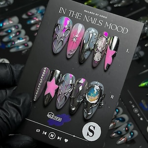 May include: A set of long, almond-shaped artificial nails with various designs. The nails feature silver, pink, and gray colors, with embellishments like stars, rhinestones, and abstract patterns. The card reads "IN THE NAILS MOOD".