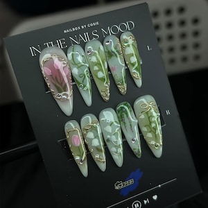 May include: A set of ten long, almond-shaped press-on nails. Each nail features a white base with floral designs in pink, green, and gold accents. The text "IN THE NAILS MOOD" is visible on the black card.