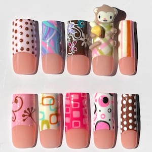 May include: A set of ten artificial nails with various designs. The nails feature a pink base with white, brown, pink, and orange patterns, including polka dots, swirls, and geometric shapes. A small monkey figurine is attached to one nail.