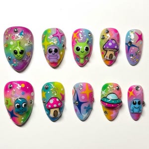May include: A set of ten almond-shaped, colorful press-on nails. Each nail features a unique design with alien and mushroom motifs, adorned with glitter and star accents. The color palette includes pink, green, blue, and yellow.