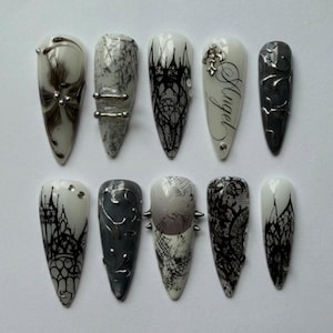 May include: A set of ten almond-shaped press-on nails featuring a monochromatic palette of black, white, and gray. Designs include intricate black lace patterns, silver metallic accents, small rhinestones, raised textures, and the word Angel in script.