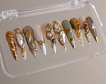 May include: A set of ten long, almond-shaped artificial nails in a clear plastic case. Each nail features a unique design with gold accents, rhinestones, and various colors, including white, silver, and muted green. The nails are designed for a fashionable manicure.