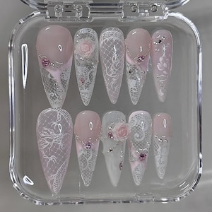 May include: A set of ten artificial nails in a clear plastic case. The nails feature a pink and white design with lace, rhinestones, and floral accents. The nails are long and pointed, with a glossy finish.