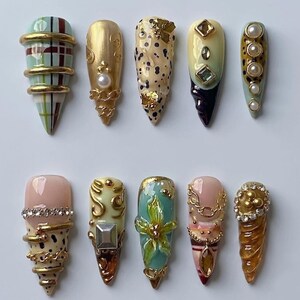 Könnte beinhalten: Ein Set aus zehn künstlichen Fingernägeln mit verschiedenen Designs, darunter Karomuster, goldene Metallic-Akzente, Perlenverzierungen, Leopardenmuster, Strasssteine und florale Motive. Jeder Nagel ist spitz geformt und hat komplexe 3D-Texturen.