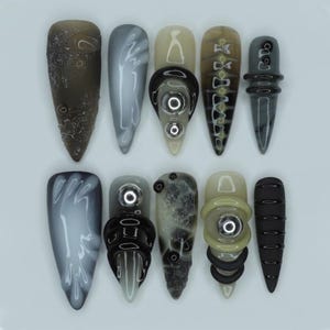 May include: A set of ten artificial nails, each with a long, pointed shape. The nails display a range of colours, such as grey, black, and beige, and are decorated with detailed patterns and embellishments. Some nails have a textured look, while others are smooth and glossy.