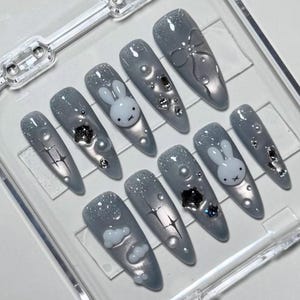 May include: A set of gray, long, almond-shaped press-on nails. Each nail features a unique design, including a white bunny, silver glitter, and various embellishments like pearls and crystals. The nails are arranged in a clear plastic container.