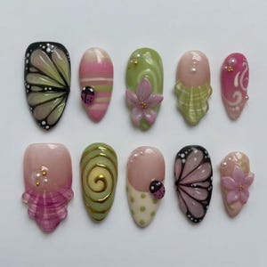 May include: A set of ten artificial nails with various designs. The nails feature butterfly wings, ladybugs, floral accents, and abstract patterns in shades of pink, green, and white. Some nails have 3D embellishments.