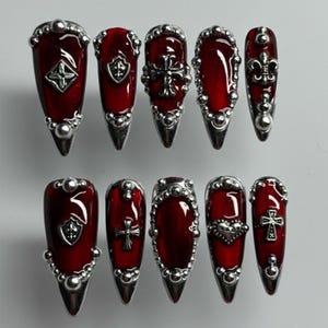 May include: A set of ten long, pointed press-on nails. Each nail has a deep red base with silver embellishments. Designs include crosses, shields, and fleur-de-lis, creating a gothic aesthetic.