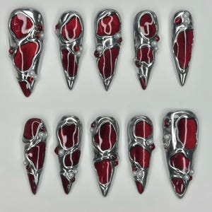 May include: A set of ten silver-toned nail covers with a deep red, textured interior. Each cover is adorned with small, pearl-like accents. The covers have a pointed, elongated shape, and are arranged in two rows of five.
