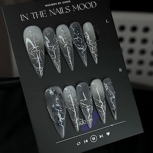Può includere: Un set di unghie finte lunghe e appuntite con un design marmorizzato grigio e bianco. Ogni unghia presenta un motivo astratto color argento unico. Le unghie sono esposte su una scheda nera con il testo "IN THE NAILS MOOD" in alto.