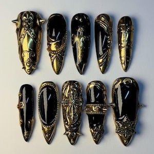 May include: A set of ten black and gold press-on nails. Each nail features a glossy black base with ornate gold embellishments, including spikes, filigree, and sculpted designs. The nails vary in shape and design, offering a unique, bold aesthetic.