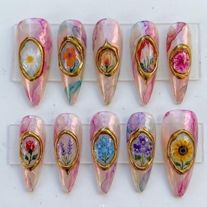 May include: Ten press-on nails featuring a translucent pink and purple marbled base. Each nail is adorned with a central, hand-painted floral motif encased in a raised, textured gold-colored oval frame. The flowers include daisies, roses, sunflowers, and lavender.