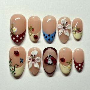 May include: A set of ten artificial nails with various designs. The nails feature ladybugs, flowers, polka dots, and other nature-inspired elements. The base color is a nude pink, with accents in red, white, blue, and green.