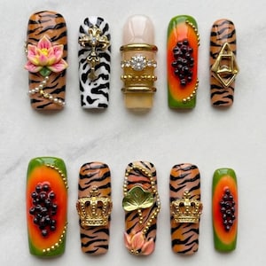 May include: A collection of ten artificial nails with various designs. The nails feature tiger stripes, fruit, floral, and jeweled accents. Some nails have gold crowns, pearls, and a cross. The color palette includes orange, green, black, white, and gold.