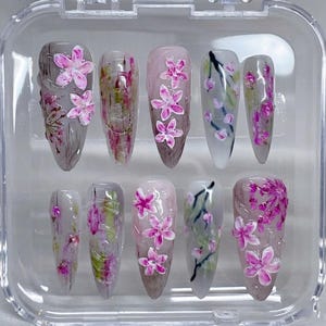 May include: A set of ten artificial nails with a floral design. The nails feature a variety of pink and white flowers, with green and brown accents. The nails are in a clear plastic case.