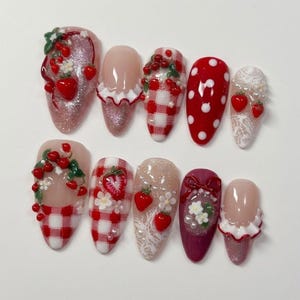 May include: A set of ten press-on nails featuring various designs including red strawberries, white flowers, red and white gingham patterns, red polka dots, white lace textures, and small white ruffles on a glossy, glittery, and solid colored base.