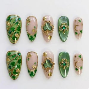 Könnte beinhalten: Ein Set aus zehn künstlichen Nägeln mit St. Patrick's Day-Motiv. Die Nägel sind in Grün-, Gold- und Nude-Tönen gehalten und mit Kleeblättern und Edelsteinen verziert. Einige Nägel haben einen goldenen Umriss, andere ein goldenes Kleeblatt-Design.