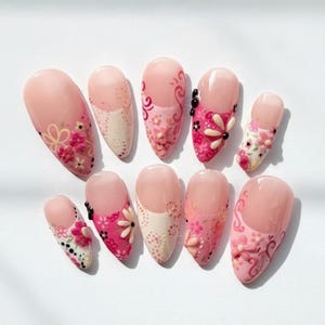 May include: A set of ten artificial nails, each with a unique design. The nails feature a light pink base with various floral patterns in shades of pink, white, and black. Some nails have 3D flower accents, while others have painted designs.