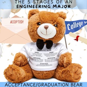 Op de afbeelding: Een bruine teddybeer die een wit T-shirt draagt met de tekst "The 5 Stages of an Engineering Major" en een zwarte strik. De tekst op het T-shirt beschrijft de stadia van ontkenning, woede, onderhandelen, depressie en acceptatie. De beer zit op een wit oppervlak met een blauw-witte afstudeerhoed en een blauw-witte banner met de tekst "College" op de achtergrond.