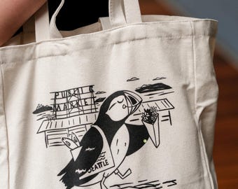 May include: A cream-coloured tote bag with a black and white illustration of a puffin carrying flowers. The bag features the text "Pike Place Market Seattle" and a stylized pier and water scene.