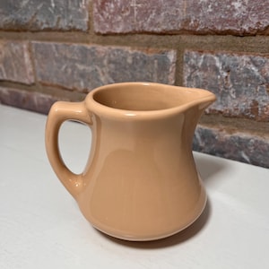 May include: A small, light brown ceramic creamer jug with a handle. The jug is sitting on a white surface.