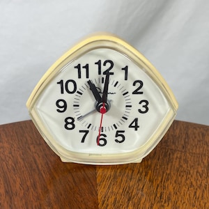 May include: A vintage, cream-colored alarm clock with a white face and black numerals. The clock has a red second hand and a geometric, trapezoid-like shape. The clock is sitting on a wooden surface.