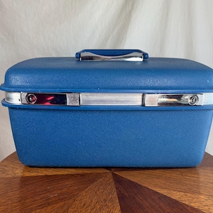 Pode incluir: Uma mala vintage azul de lados rígidos com uma alça e acabamentos prateados. A mala tem formato retangular com cantos arredondados e uma superfície texturizada. As travas e a alça de metal são brilhantes. A mala está em cima de uma mesa de madeira.