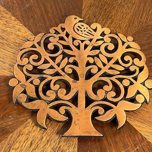 May include: A copper-coloured metal trivet shaped like a tree, with a bird at the top. The tree design has detailed cut-out leaves and branches. The trivet is round and rests on a wooden surface. The trivet's diameter is approximately 18 cm.