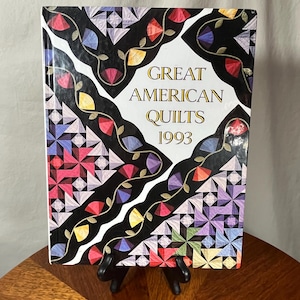 May include: A hardcover book titled "Great American Quilts 1993" is displayed on a black stand. The cover features a black background with colorful quilt block designs and the title in gold lettering.