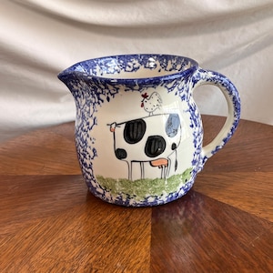 May include: A ceramic pitcher with a blue and white speckled rim and handle. The pitcher features a hand-painted design of a cow with black spots and a chicken on its back. The background is white with blue accents.