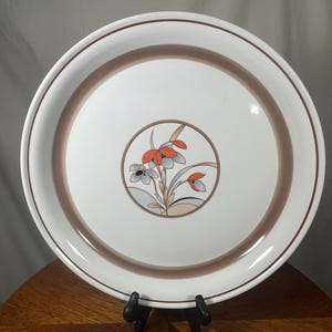 May include: White ceramic dinner plate with a brown rim and a floral design in the center. The design features orange, gray, and beige flowers and leaves within a circular border. The plate is displayed on a wooden surface.