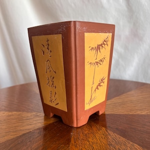 Könnte beinhalten: Ein quadratischer, terrakottafarbener Blumentopf mit Bambusdesign und chinesischen Schriftzeichen. Der Topf hat einen hellbraunen Hintergrund mit einer Bambusillustration und Text. Der Topf steht auf einer Holzoberfläche.