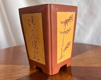 May include: A square, terracotta-colored planter with a bamboo design and Chinese characters. The planter has a light brown background with a bamboo illustration and text. The planter is sitting on a wooden surface.