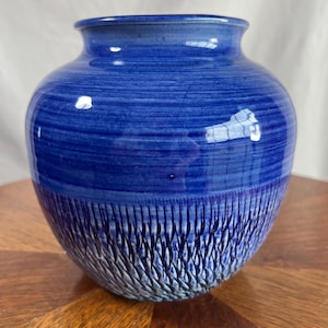 Puede incluir: Un jarrón de cerámica azul con un diseño texturizado y tallado en la mitad inferior. La parte superior tiene un patrón de rayas horizontales suaves. El jarrón es de color azul oscuro y tiene forma redondeada.