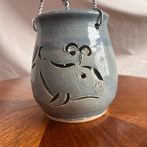 Puede incluir: Un portavelas colgante de cerámica azul con un diseño de ballena recortada. El portavelas tiene una cadena de metal para colgar.