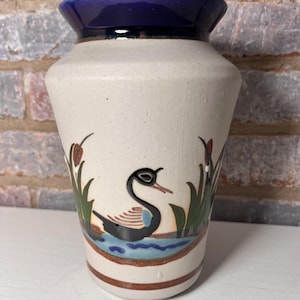 Puede incluir: Un jarrón de cerámica blanco con un borde azul y un diseño pintado de un cisne negro nadando en un estanque azul con juncos verdes.