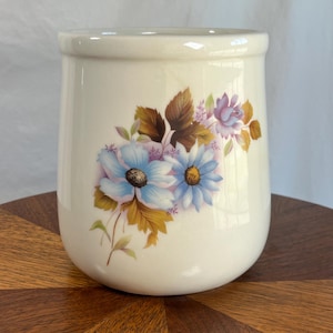 May include: Cream-colored ceramic utensil holder with a floral design. The flowers are light blue and purple with brown and yellow leaves. The holder is cylindrical with a slightly flared top.