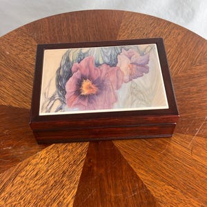 Könnte beinhalten: Eine rechteckige Holzbox mit dunkelbraunem Finish. Der Deckel zeigt ein Blumendesign mit lila und rosa Blüten. Die Box steht auf einem Holztisch mit gemusterter Oberfläche. Die Box wird wahrscheinlich zur Aufbewahrung von Schmuck oder kleinen Gegenständen verwendet.