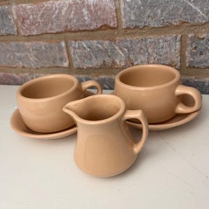 May include: A set of three pale pink ceramic teacups and a creamer. The cups have a simple design and are perfect for a casual tea party.