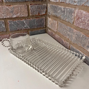 Può includere: Un vassoio da portata in vetro trasparente con un bordo smerlato e una piccola tazza di vetro con manico. Il vassoio ha un design a coste ed è perfetto per servire spuntini o bevande.
