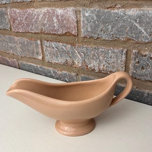 May include: A light pink ceramic gravy boat with a handle and a pedestal base.