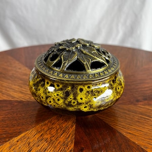 May include: A round ceramic bowl with a glossy yellow and black speckled glaze, topped with a removable dark bronze-toned metal lid featuring an intricate openwork floral and leaf pattern and a decorative triangular border around the rim.
