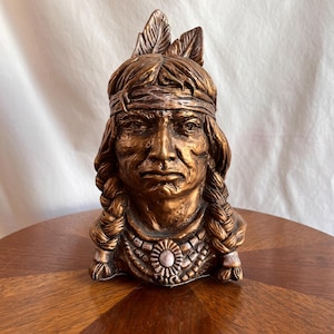 Puede incluir: Busto de color bronce de un nativo americano. La escultura presenta rasgos faciales detallados, cabello largo trenzado y un tocado de plumas. Un collar de cuentas con un colgante central adorna el pecho. El busto se exhibe sobre una mesa de madera.
