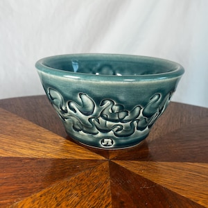 May include: A teal ceramic bowl with a glossy, crackled glaze finish. The exterior features a raised, swirling, wave-like pattern encircling the body. Near the base, a small, stamped maker's mark is visible, consisting of the letters J and R.