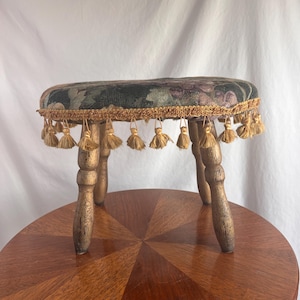 May include: A small, antique wooden stool with a floral patterned seat in shades of green, purple, and black. The seat is trimmed with gold-colored tassels. The stool has four turned legs and is displayed on a round wooden table.
