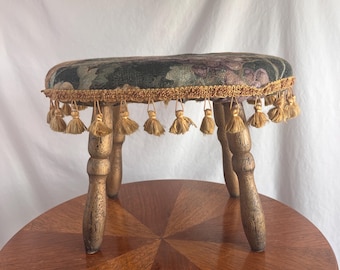 May include: A small, antique wooden stool with a floral patterned seat in shades of green, purple, and black. The seat is trimmed with gold-colored tassels. The stool has four turned legs and is displayed on a round wooden table.